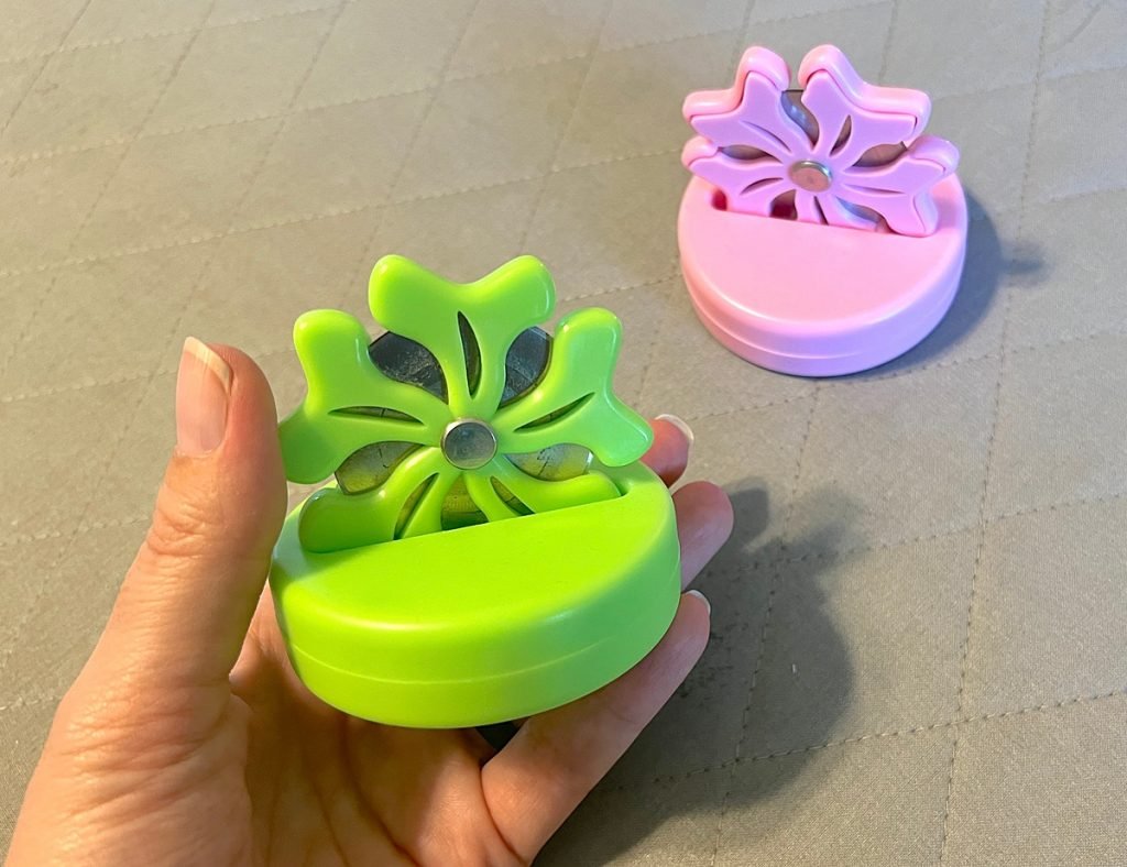 Two flower shaped thread cutters. One is pink, the other is green and shown in a woman's hand.
