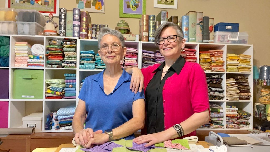 Adult mother and daughter smiling in front of a collection of colorful, folded fabrics.